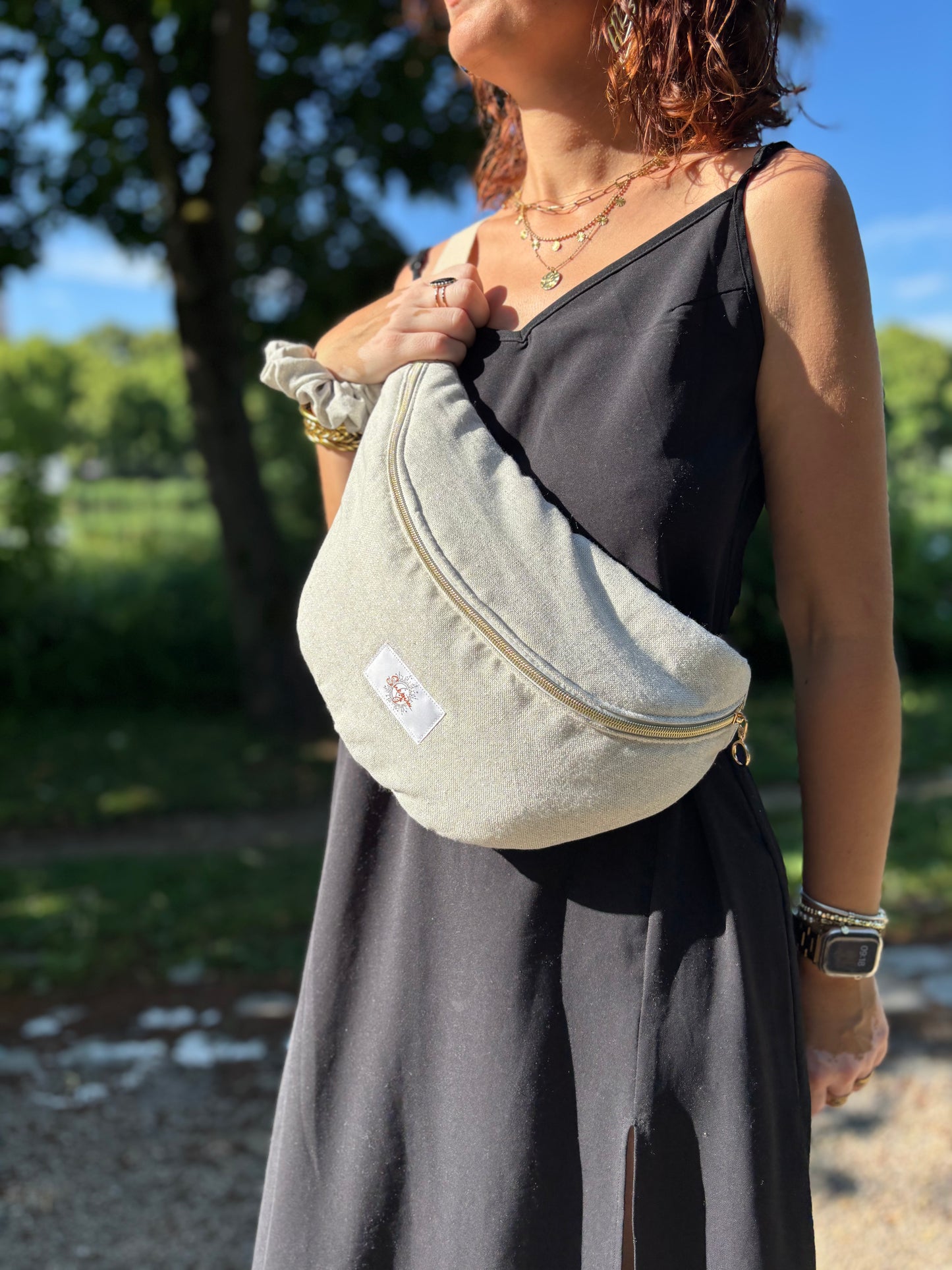 Banane beige pailleté portée en bandoulière par une femme dans un parc ensoleillé, symbole de liberté et de style au quotidien. Ce modèle allie audace et douceur, parfait pour garder les mains libres avec élégance. Confectionnée à la main avec un tissu jacquard de qualité, elle ne se déforme pas et conserve sa tenue. Idéale pour accompagner les journées actives avec une touche de féminité bohème.