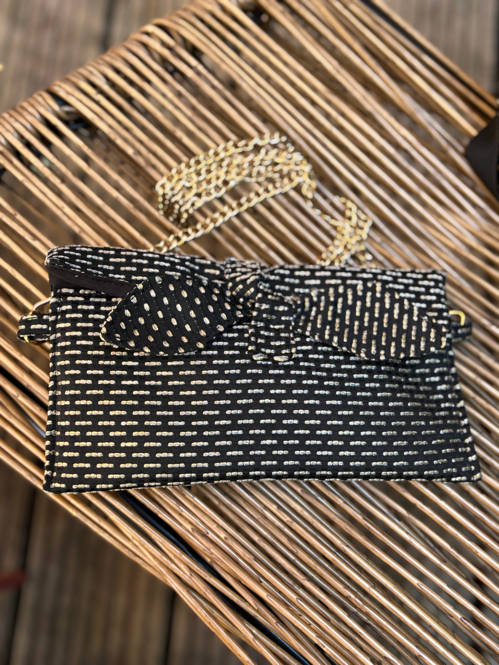 Trousse noire Alma de SueGiu posée à plat, avec sa forme rectangulaire et ses détails sobres et élégants. Tissu texturé noir profond, fermeture éclair dorée, posé sur une chaise en rotin